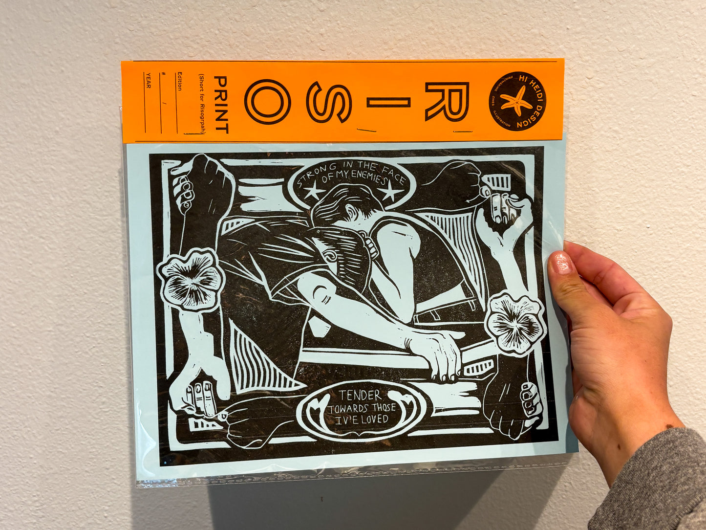 Hand holding a print with black and white design on an orange border against a beige wall.