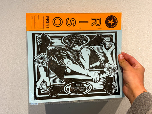 Hand holding a print with black and white design on an orange border against a beige wall.