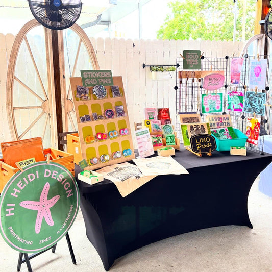 Image is of Hi Heidi Designs Art Market Setup inclduding risograph prints, linocut prints, and handmade pins and stickers